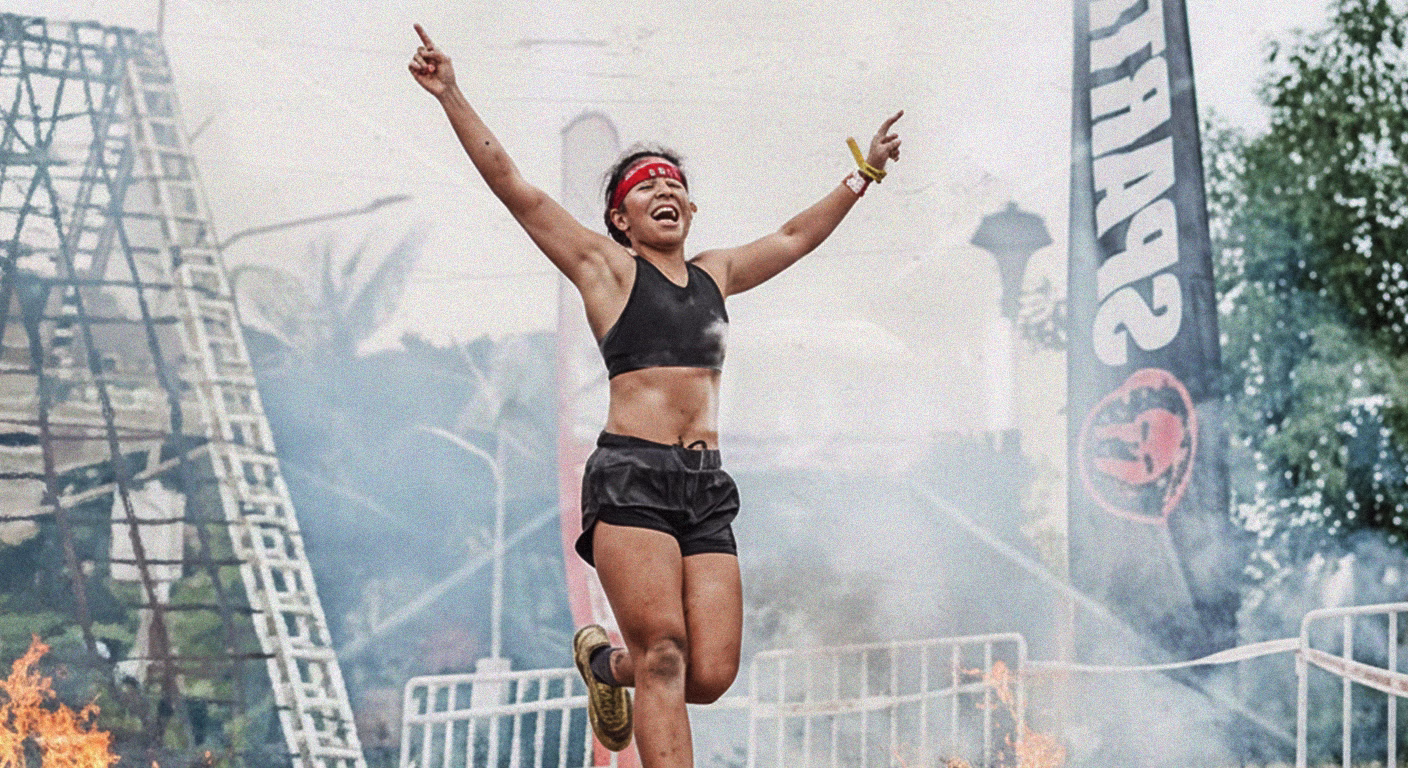Alyana Chavez, wearing a red headband and athletic wear, triumphantly leaps with arms raised at a Spartan Race obstacle course, with a fire pit and the Spartan logo in the background. - heatseekerproject.com